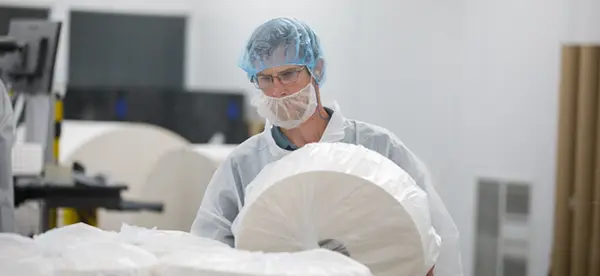 Worker in a hairnet, beard cover, safety glasses and white protective gown inspecting a large roll of white nonwoven material in a clean manufacturing facility.